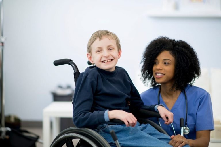 Smiling child in wheelchair with supportive nurse beside him.