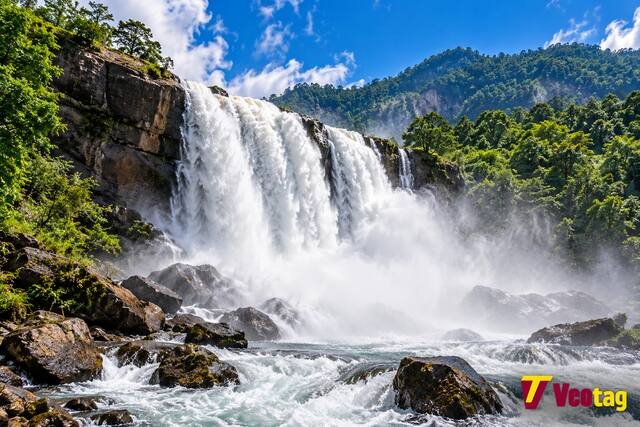 High-force waterfall cascading over rocky cliffs into a powerful river surrounded by lush green forest.