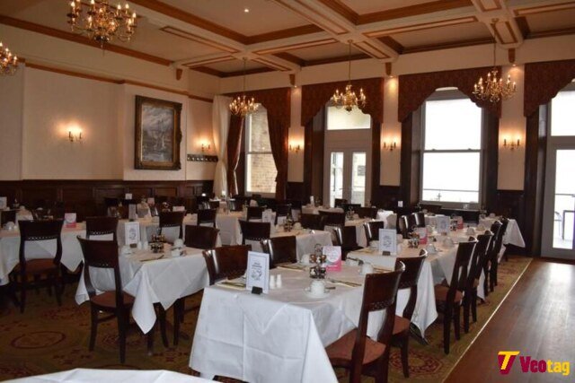 Elegant restaurant dining room with white tablecloths, wooden chairs, chandeliers, and large windows.