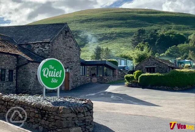 Entrance to The Quiet Site with traditional stone buildings, a green sign, and scenic hills in the Lake District.